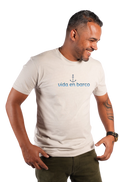 A man wearing a light beige crewneck t-shirt with a blue anchor graphic and the text 'vida en barco' printed on the chest, paired with olive green pants, set against a plain white background.