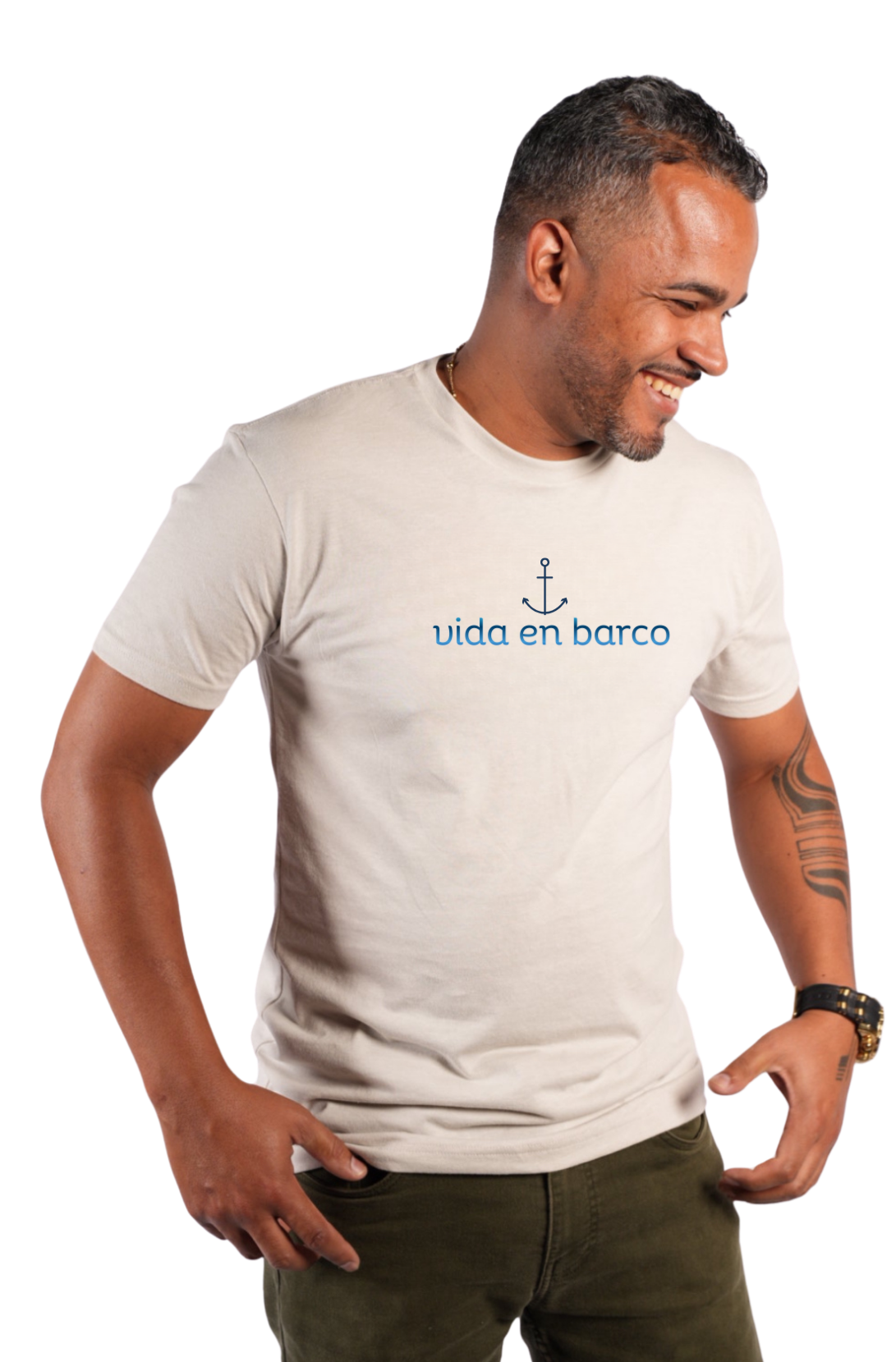 A man wearing a light beige crewneck t-shirt with a blue anchor graphic and the text 'vida en barco' printed on the chest, paired with olive green pants, set against a plain white background.
