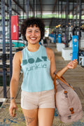 A smiling woman with curly dark hair stands on a boat dock wearing a light blue tank top with 'BOAT junkie' printed in dark teal, paired with beige shorts and carrying a large beige weekend bag.