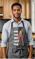 A man wearing a dark gray kitchen apron with the text 'WHO SAID MEN CAN'T COOK' and a red-handled tool graphic, standing in a kitchen with wooden cabinets.