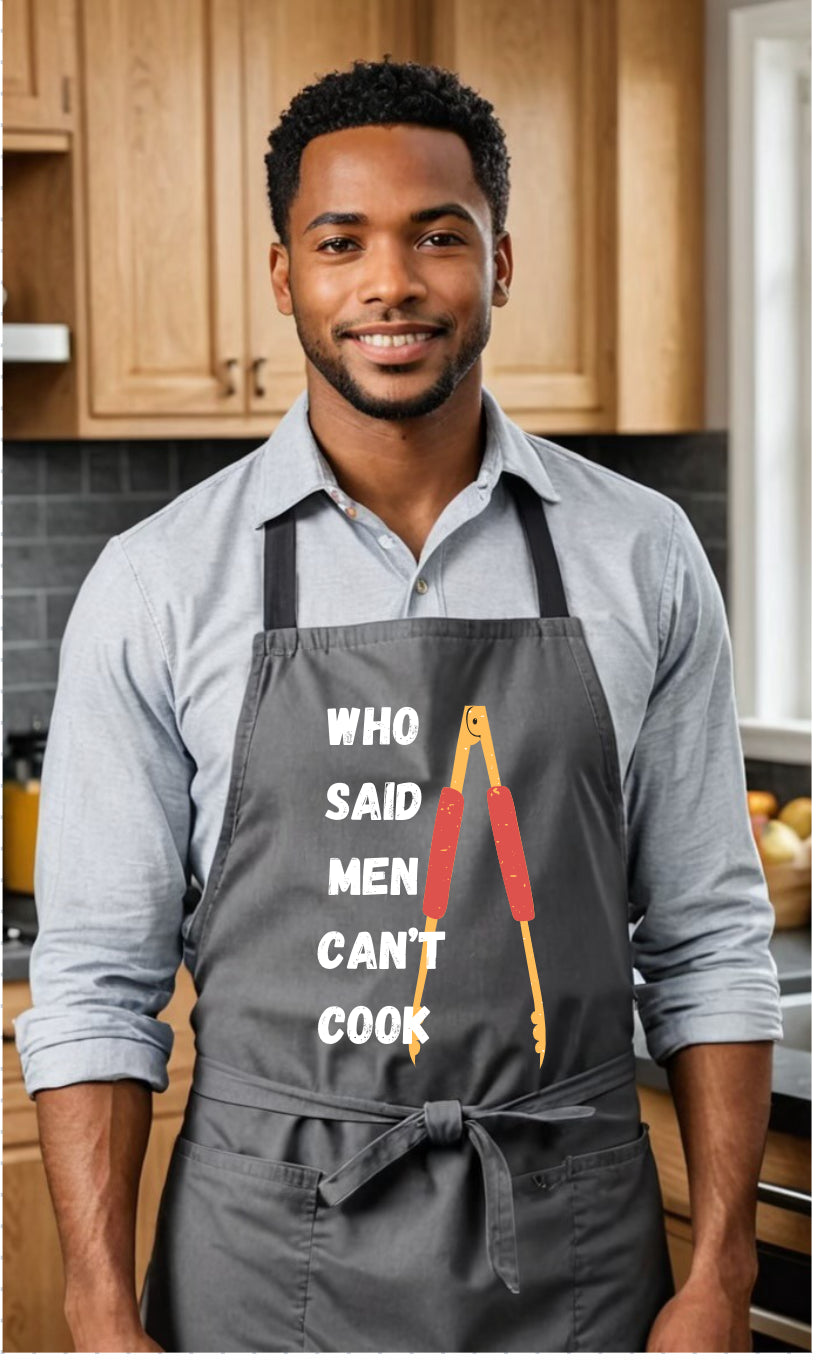 A man wearing a dark gray kitchen apron with the text 'WHO SAID MEN CAN'T COOK' and a red-handled tool graphic, standing in a kitchen with wooden cabinets.
