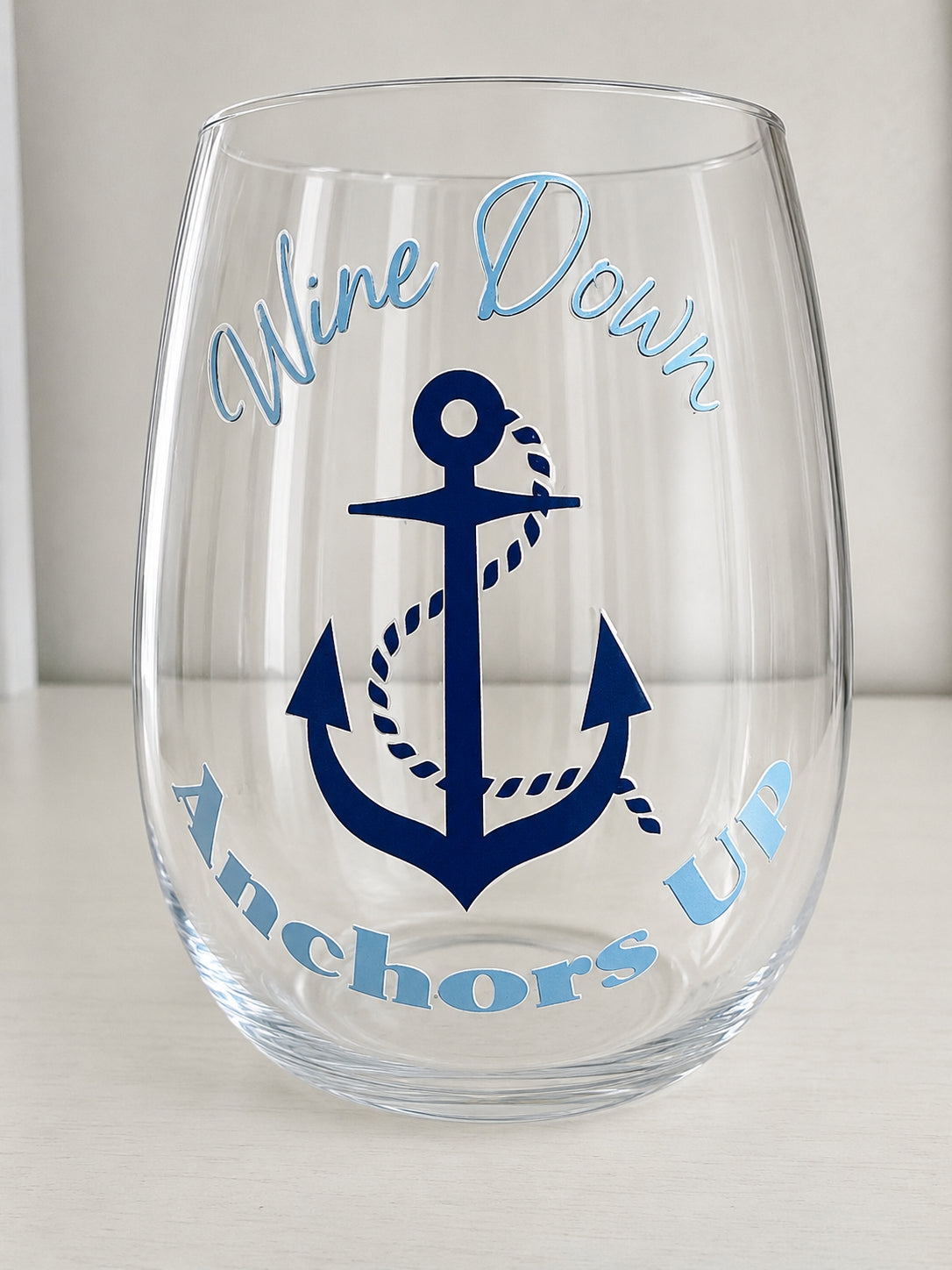 A clear shatterproof stemless wine glass with a navy blue anchor graphic and the words 'Wine Down Anchors Up' in light blue script, shown on a light neutral background.
