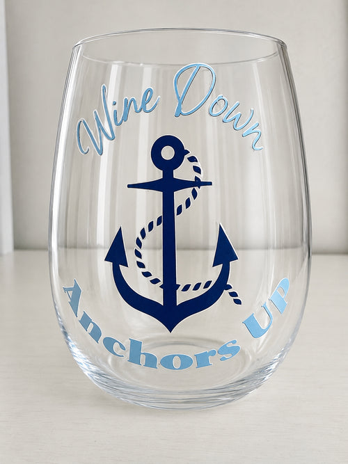 A clear shatterproof stemless wine glass with a navy blue anchor graphic and the words 'Wine Down Anchors Up' in light blue script, shown on a light neutral background.