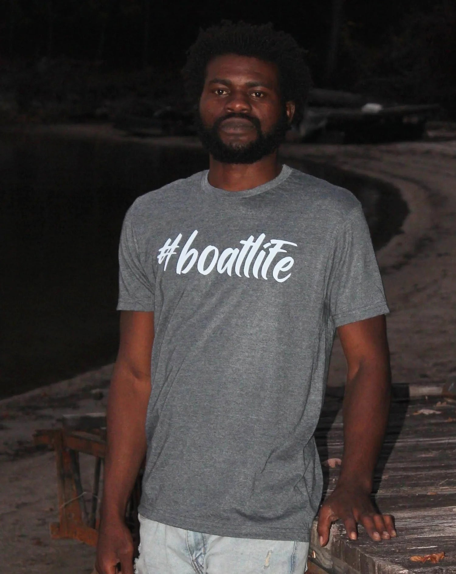A man wearing a heather gray t-shirt with the white text '#boatlite' printed across the chest, standing outdoors on a wooden dock at night.