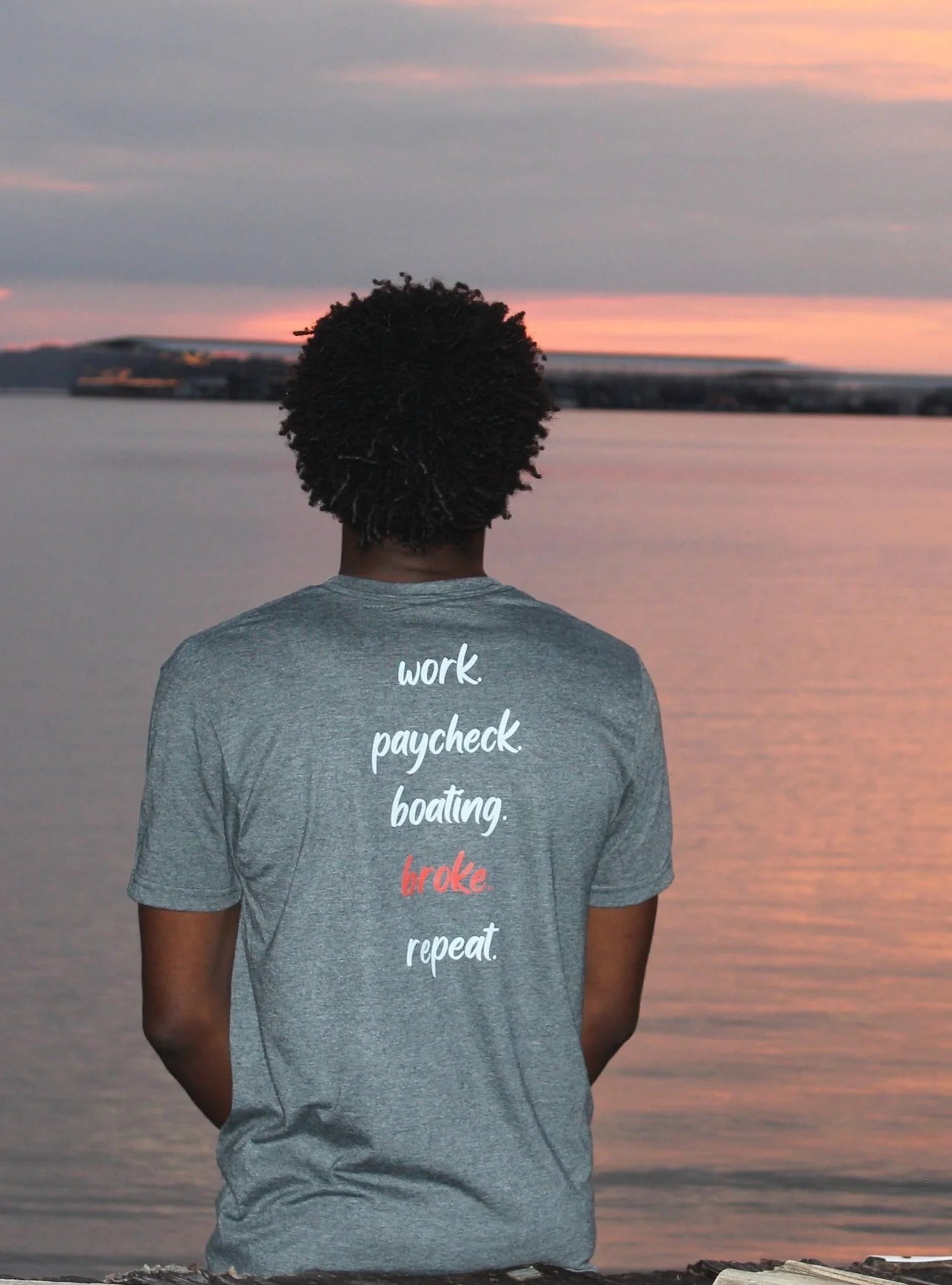 A man seen from behind wearing a heather gray t-shirt with a humorous quote printed on the back, set against a scenic sunset over water.